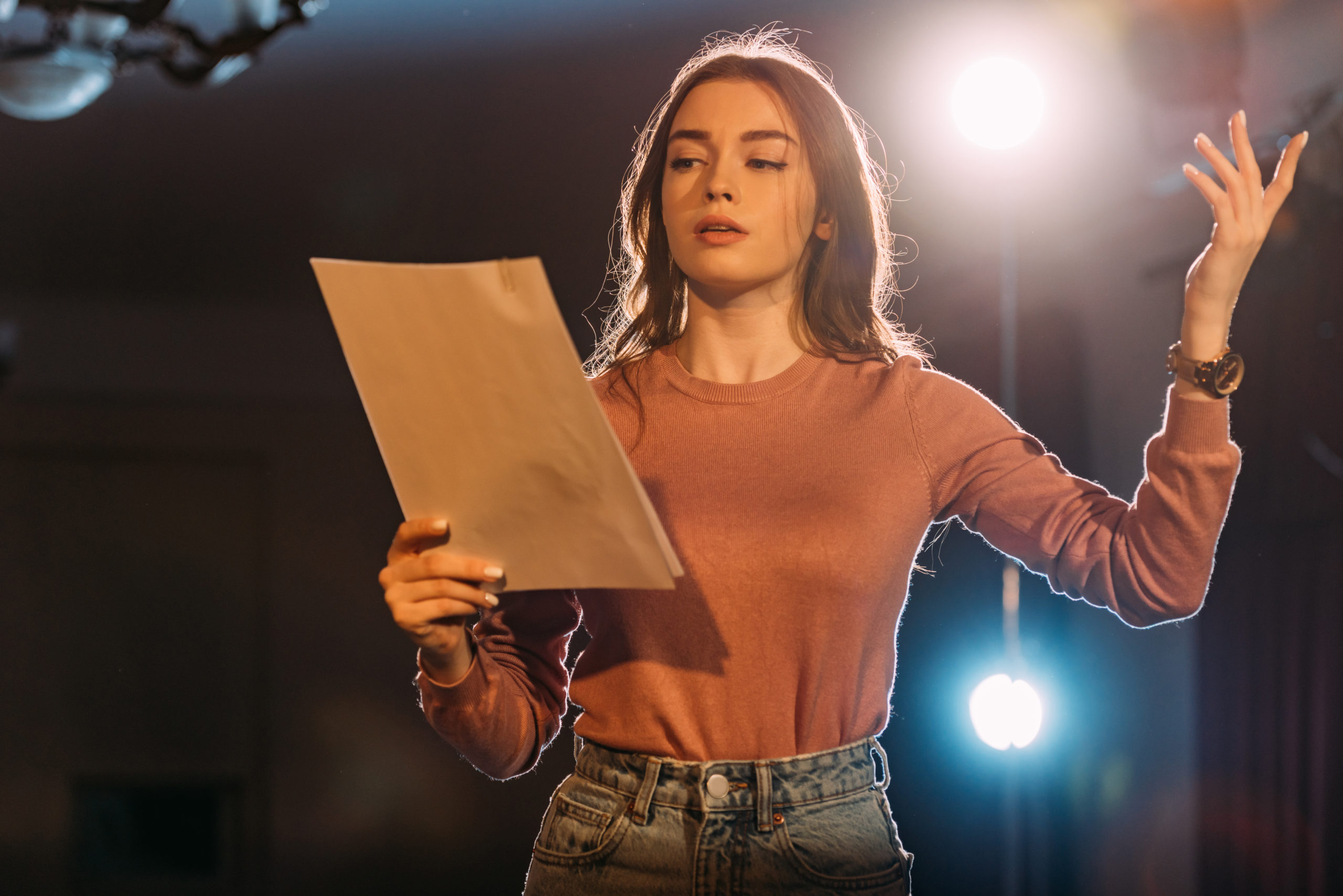 A woman standing, reading from a script and holding her hand aloft.