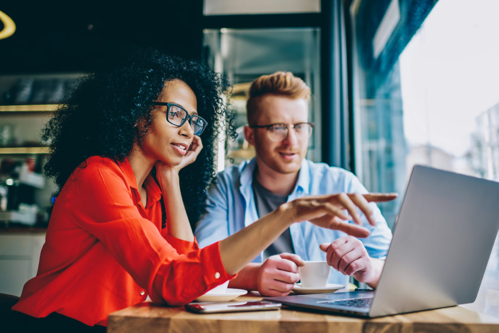 Stock photo of two colleagues, sitting in front of a laptop. The person on the left is pointing out something to the person on the right. They are next to a bright window in a modern office.